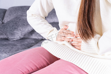 疼痛摄影照片_坐在家里的沙发上患有强烈腹痛或月经的年轻女性。