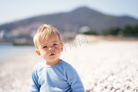 严肃的孩子坐在海边的卵石滩上，背靠群山。