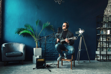 男歌手摄影照片_男歌手坐在凳子上，戴着耳机，吉他在家庭录音室录制曲目