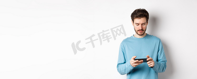 男模在智能手机上玩电子游戏，看着手机屏幕很严肃，站在白色背景上的运动衫