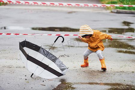 雨后穿着黄色防水斗篷、靴子和雨伞在户外玩耍的孩子靠近保护带