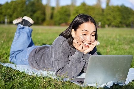 美丽女孩躺在公园里，惊讶地看着笔记本电脑、观看视频、在户外放松时与朋友聊天的肖像