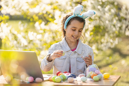 快乐的少女戴着兔耳朵在花园里用笔记本电脑画复活节彩蛋
