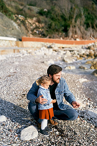 微笑的爸爸蹲在小女孩旁边，递给她从水边的岩石海滩上捡来的鹅卵石。