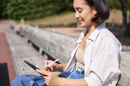 亚洲年轻女性在公园里呼吸新鲜空气的画像，坐在平板电脑和数字笔旁，开心地笑着