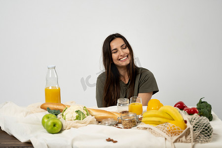 穿着绿色 T 恤的快乐女人在家里的厨房里打开购物网状生态袋，里面装着健康的蔬菜、水果、面包、零食。