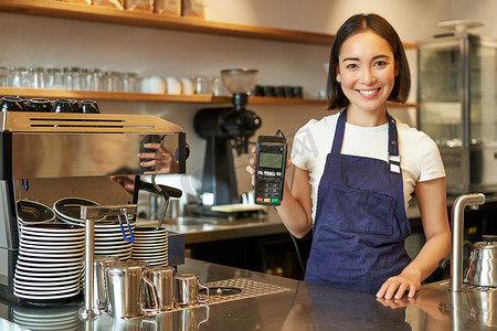 微笑的亚洲女咖啡师，围裙的咖啡馆老板，展示卡片机，支付读卡器，在她的咖啡店接受非接触式订单