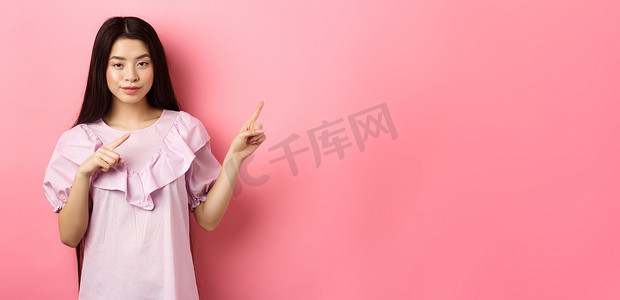 厚颜无耻的亚洲女孩展示有趣的广告，用手指指着标志，狡猾地微笑，暗示很划算，粉红色背景