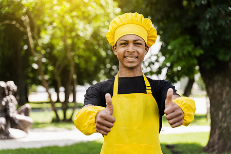 非洲黑人少年厨师戴着厨师帽竖起大拇指，身穿黄色围裙制服，为面包店做面团。