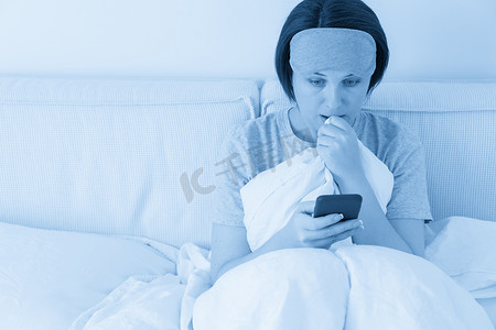 戴着睡眠面具的女人坐在铺着羽绒被的床上，用智能手机发短信
