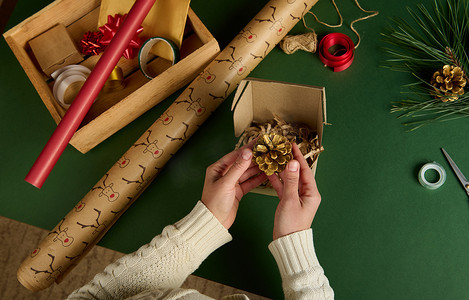 顶视图女人的手把金松果放在工艺纸板箱里，同时包装圣诞礼物。