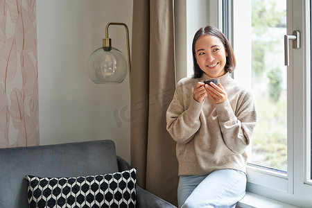喝咖啡表情摄影照片_美丽的亚洲年轻女性坐在窗边喝咖啡，拿着浓咖啡杯，面带轻松、微笑的表情看着外面