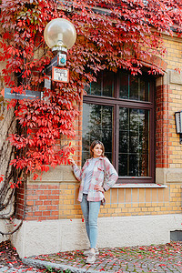红背景2022摄影照片_一个美丽的女孩站在欧洲老房子的窗户的背景下，与秋天的飘红交织在一起。