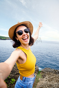 微笑天使摄影照片_快乐和微笑的年轻女性旅行者在暑假期间在海边位置用手机自拍。