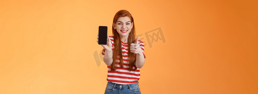 快乐满意的红发迷人女孩欢呼竖起大拇指，推荐智能手机应用程序，显示手机屏幕，介绍设备，批准好衣服秀在线商店手机，橙色背景