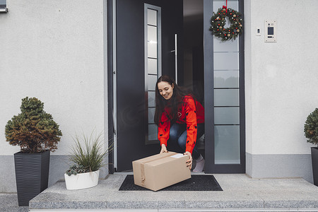 身穿红色圣诞毛衣的年轻女子捡起留在前门的纸箱邮递员