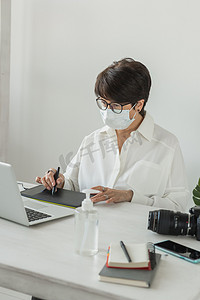 医用面罩平面设计师或摄影师在现代办公室工作时使用数字图形平板电脑的中年妇女，坐在现代工作区的专业女性修饰师，通用设计平板电脑和附近的相机。 