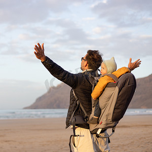 日落时，年轻的父亲在西班牙兰萨罗特岛 Famara 多风的沙滩上，一边享受纯净的大自然，一边背着他的婴儿男婴儿子背着背包，举起双手向天空。