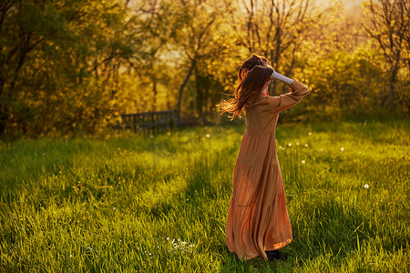 一位留着长发的苗条女性背对着镜头站在田野里，被落日的光芒照亮，愉快地摆出姿势享受温暖的天气和休息