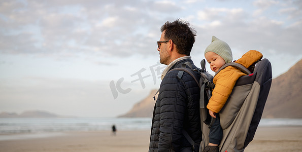 日落时分，父亲在西班牙兰萨罗特岛法马拉多风的沙滩上，背着他的婴儿男孩儿子在背包里享受纯净的大自然。