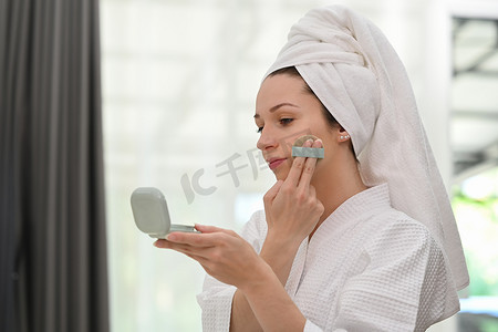 穿着浴袍、带垫粉扑和镜子的漂亮白人女性，在脸上涂上化妆粉底