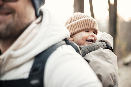 连衣裙子摄影照片_运动型父亲背着他的小儿子穿着冬季连身衣和帽子，背着背包在秋天的森林里徒步旅行。