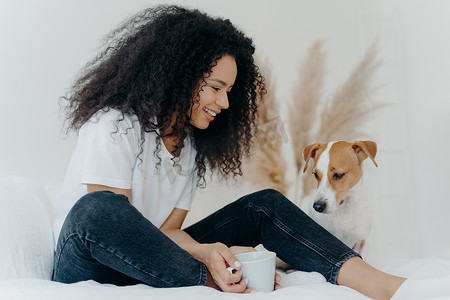可爱的卷发女狗主人的照片看着动物微笑，拿着一杯茶，坐在白色宽敞卧室的床上，表达对动物的爱。