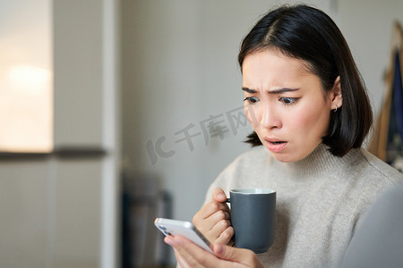 手拿杯子摄影照片_手拿杯子的亚洲女孩特写，看着手机屏幕感到震惊，阅读有关智能手机应用程序的新闻