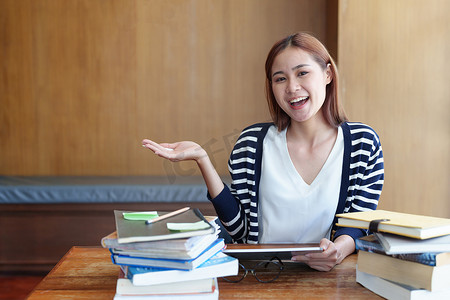 一幅年轻亚洲女性的画像，她在图书馆使用平板电脑在线学习时面带微笑，并用手势表达想法