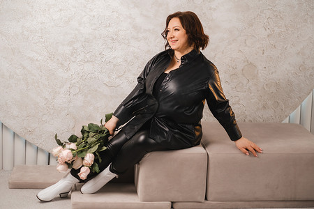 一位穿着黑色皮衣的时尚成年女性坐在室内，手里拿着一束粉红玫瑰
