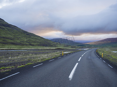 柏油路穿过空荡荡的北部景观，绿草如茵，五颜六色的山丘，水坑和日落戏剧性的天空，通往冰岛西部高地的山脉，复制空间