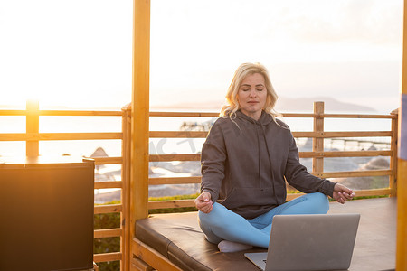 一位平静的女士坐在海边的日光躺椅上，在电脑前工作时分心，冥想并消除负面情绪，将远程工作与休息结合起来。