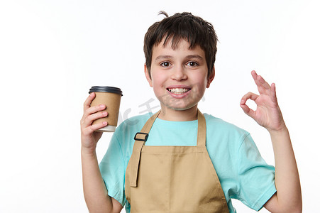 聖保路标志摄影照片_孤立的肖像白色背景：快乐的十几岁男孩酒保展示 OK 标志，用生态纸杯出售外卖咖啡