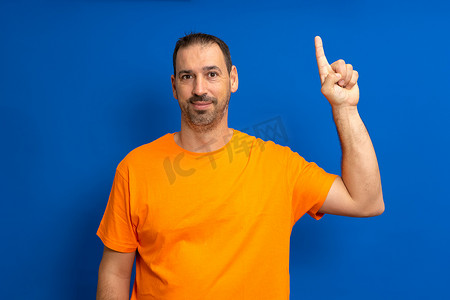 40 多岁有见地、聪明、积极主动的白人男子，身穿橙色 T 恤，举着食指，提出了与蓝色背景工作室肖像隔离的伟大新想法。