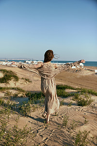 一位身穿米色连衣裙的优雅女性背对着镜头，在远处可见的大海背景下走在沙滩上