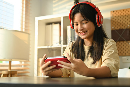 电子游戏摄影照片_戴着耳机在便携式游戏机上玩电子游戏的快乐亚洲少女