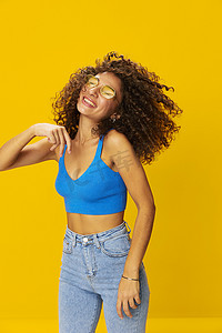 黄色背景上身穿蓝色 T 恤、卷发爆炸头的女人，戴着黄色太阳镜、手标舞动飞扬的头发，看着镜头，微笑着牙齿和幸福，复制空间