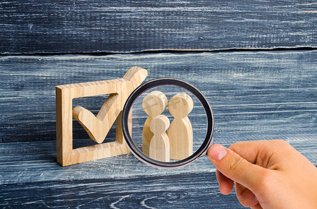 人形符号摄影照片_放大镜正在观察三个木制人形站在盒子里的一个勾号旁边。