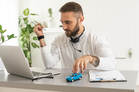 汽车保险在线概念：一名男子手持带清单、玩具车、桌上笔的智能手机，并使用笔记本电脑在线检查事故情况。