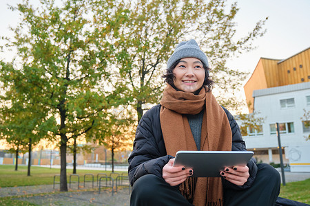 天气app摄影照片_穿着暖和衣服的亚洲女孩的画像，坐在长椅上，拿着数字平板电脑和图形笔，开心地笑着，在寒冷的天气里在户外画画