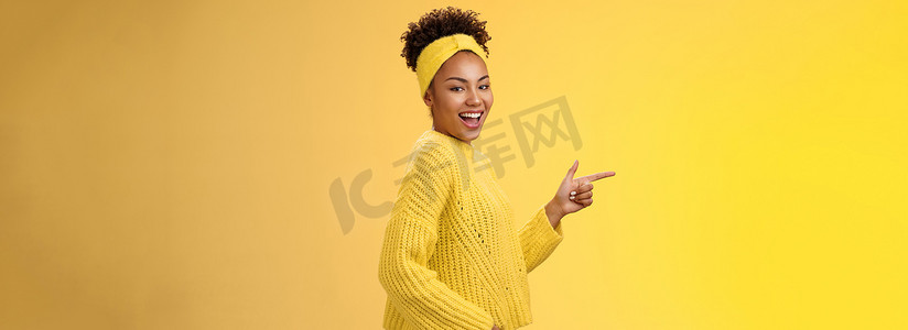 时髦可爱的女性非洲裔美国女孩 20 多岁，头带毛衣，面带微笑，面带轻浮的表情，相机转动站立的侧面，指向左侧，介绍令人敬畏的新产品展示方式，黄色背景
