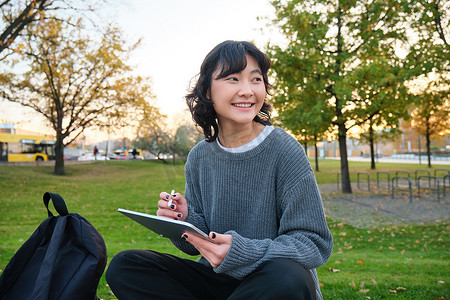 年轻的韩国女性坐在公园里，在数字平板电脑上画画，用钢笔工具在小工具上写字，环顾四周并做笔记，创作艺术品或划痕