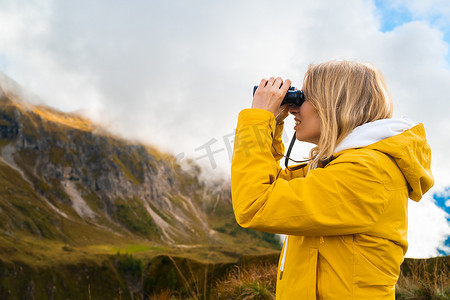 中筒摄影照片_年轻的金发女子在山中徒步旅行并通过双筒望远镜观察