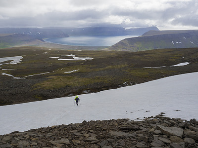 孤独的徒步旅行者背着沉重的背包走在冰岛西峡湾自然保护区 Hornstrandir 的雪地上，可以欣赏到阿达尔维克湾、海洋、山丘和陡峭的悬崖，乌云密布的天空背景