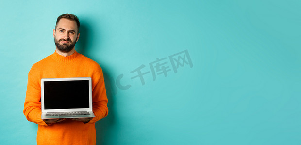 身穿橙色毛衣、留着胡子的帅哥展示笔记本电脑屏幕，展示促销，站在浅蓝色背景上，脸上露出失望和不安的表情