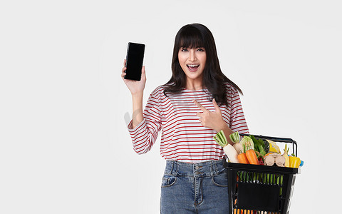 快乐的亚洲女人带着智能手机拿着装满新鲜蔬菜杂货的篮子，背景是白色的。