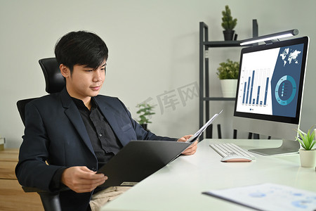 坐着的会计摄影照片_自信的亚洲男性会计顾问坐在工作站时拿着带文件的开放式办公室活页夹