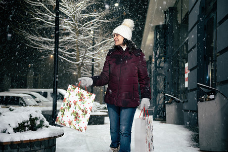 冬天购物袋摄影照片_令人愉快的年轻女子走在街上，手里拿着购物袋，手里拿着圣诞礼品盒。