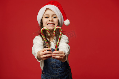 齿形边框摄影照片_细节：戴着圣诞帽的孩子的手，在相机前伸出红色背景上的红绿条纹棒棒糖
