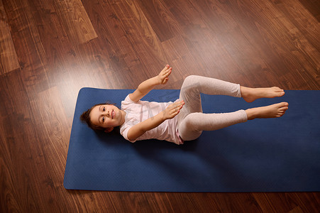 俯视可爱的小女孩在做腹肌锻炼时举起手臂，躺在蓝色的健身垫上，对着镜头微笑